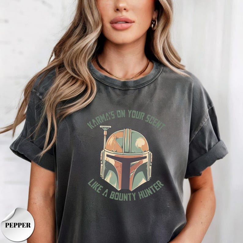May include: A charcoal gray short-sleeve t-shirt featuring a graphic of a helmet in shades of green, orange, and brown. The text "KARMA'S ON YOUR SCENT LIKE A BOUNTY HUNTER" is printed in green lettering in an arc above and below the helmet graphic.