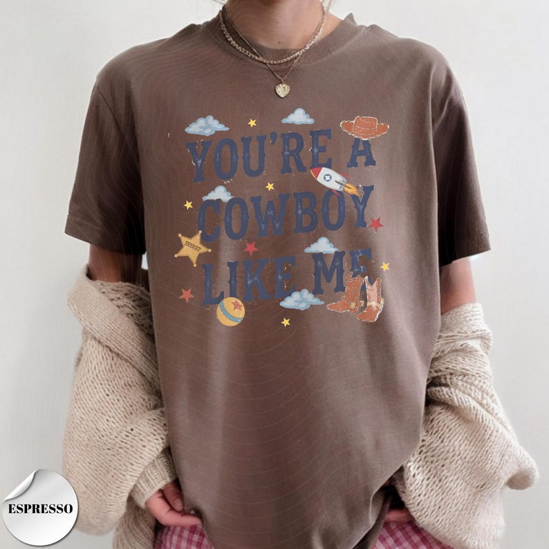 May include: A brown t-shirt featuring the blue text YOU'RE A COWBOY LIKE ME, surrounded by graphic illustrations of a cowboy hat, a rocket ship, a sheriff badge, a ball, clouds, and stars. The shirt is worn with a beige cardigan and pink plaid pants.