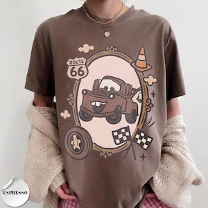 May include: A brown t-shirt featuring a graphic print of a brown tow truck character inside an oval frame. The design includes a Route 66 sign, a traffic cone, a tire, and two checkered flags. The text "ESPRESSO" appears in a small circular badge.