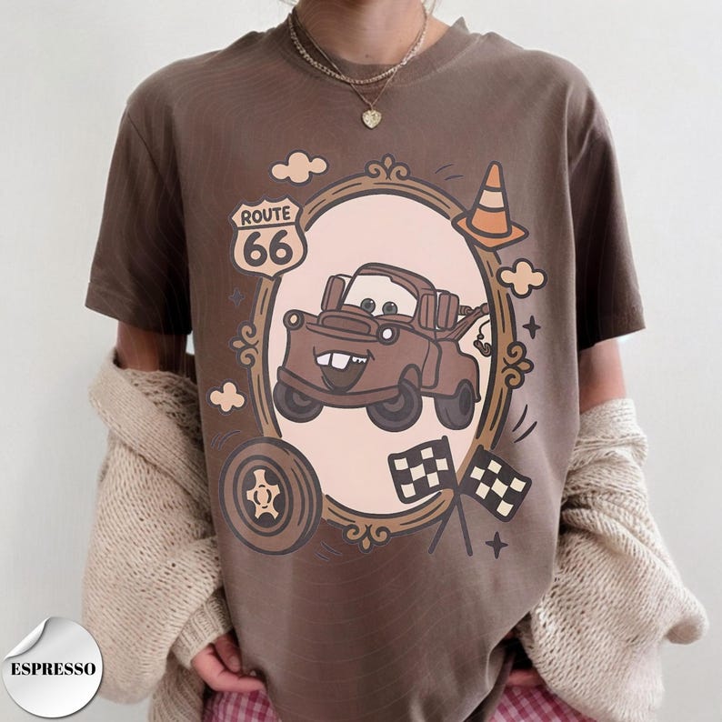 May include: A brown t-shirt featuring a graphic print of a brown tow truck character inside an oval frame. The design includes a Route 66 sign, a traffic cone, a tire, and two checkered flags. The text "ESPRESSO" appears in a small circular badge.