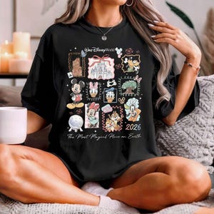 K&ouml;nnte beinhalten: Schwarzes T-Shirt mit einem Disney World-Design, das Figuren wie Micky Maus und Donald Duck sowie das Schloss von Aschenputtel zeigt. Das Shirt enth&auml;lt den Text "2026" und "The Most Magical Place on Earth."