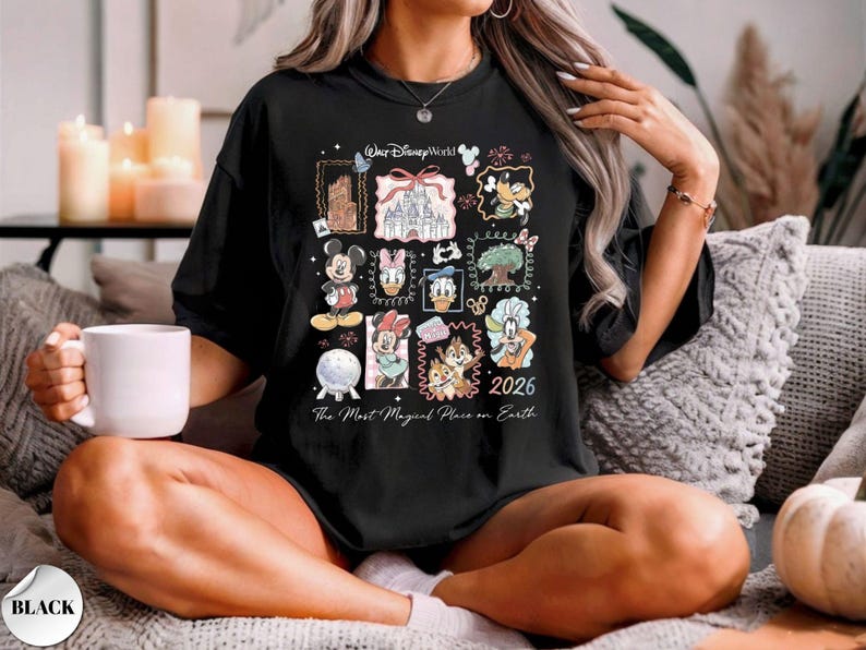 K&ouml;nnte beinhalten: Schwarzes T-Shirt mit einem Disney World-Design, das Figuren wie Micky Maus und Donald Duck sowie das Schloss von Aschenputtel zeigt. Das Shirt enth&auml;lt den Text "2026" und "The Most Magical Place on Earth."
