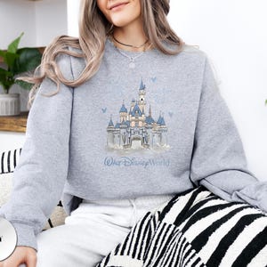 May include: A heather grey crewneck sweatshirt featuring a central graphic illustration of a blue and white castle with the text Walt Disney World printed underneath in a script font. The design includes small blue bird accents around the castle.