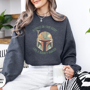 Puede incluir: Una sudadera de cuello redondo en color gris marengo jaspeado con un gr&aacute;fico de un casco estilizado en tonos verde, beige y marr&oacute;n. El texto sobre el casco dice KARMA'S ON YOUR SCENT y el texto debajo dice LIKE A BOUNTY HUNTER en verde.
