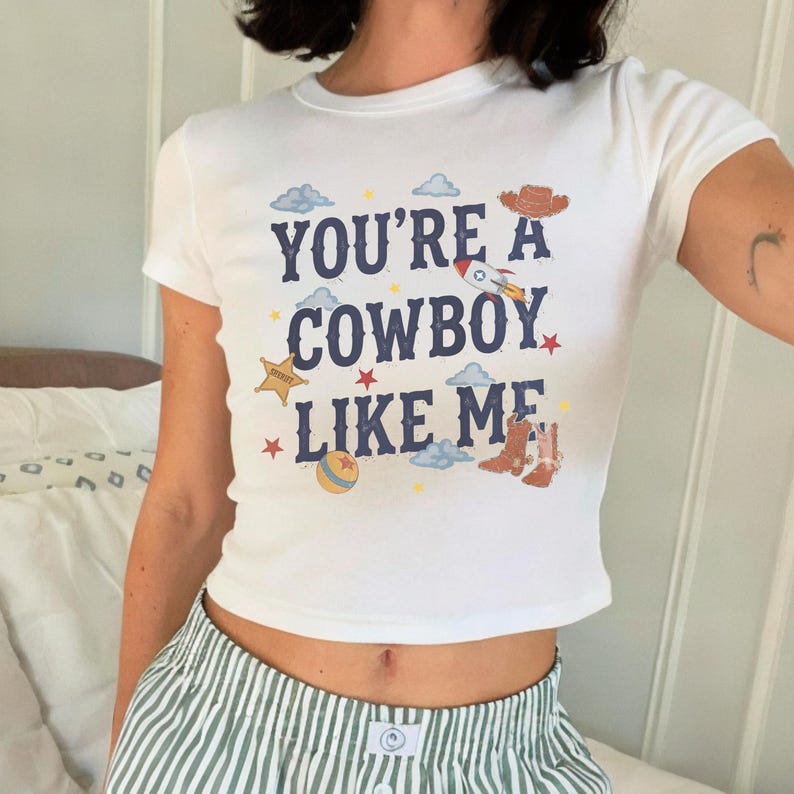 May include: A white short-sleeved t-shirt featuring the dark blue text YOU'RE A COWBOY LIKE ME. The graphic includes illustrations of a brown cowboy hat, a rocket ship, a yellow sheriff star, a colorful ball, and brown cowboy boots with clouds.