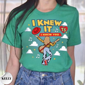 May include: A green t-shirt featuring a graphic of a person with long hair wearing a yellow plaid shirt and cow-print pants, holding a guitar. The text reads I KNEW IT and I KNEW YOU, accompanied by a red hat, a red heart with TS, clouds, and music notes.
