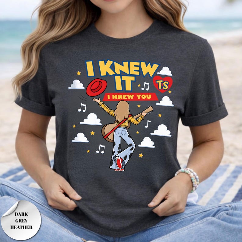 May include: A dark grey heather t-shirt featuring a graphic of a person with long hair playing a guitar, wearing a yellow shirt and cow-print pants. The text reads I KNEW IT and I KNEW YOU, accompanied by a red hat, a red heart with TS, and clouds.