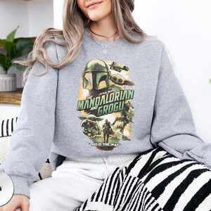 Puede incluir: Una sudadera gris de cuello redondo con un estampado gr&aacute;fico de The Mandalorian y Grogu. El dise&ntilde;o incluye el texto "THE MANDALORIAN AND GROGU" y "THIS IS THE WAY" con ilustraciones de los personajes en una paleta de colores suaves.