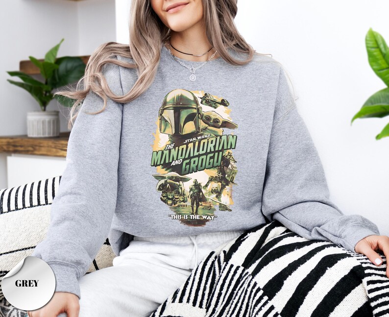 Puede incluir: Una sudadera gris de cuello redondo con un estampado gr&aacute;fico de The Mandalorian y Grogu. El dise&ntilde;o incluye el texto "THE MANDALORIAN AND GROGU" y "THIS IS THE WAY" con ilustraciones de los personajes en una paleta de colores suaves.