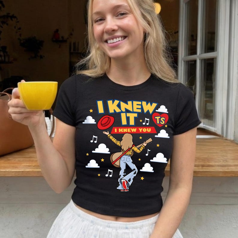 May include: A black short-sleeved t-shirt featuring a graphic print with the text I KNEW IT and I KNEW YOU. The design includes a cartoon figure with long hair playing a guitar, a red hat, a red heart with TS, musical notes, and white clouds.