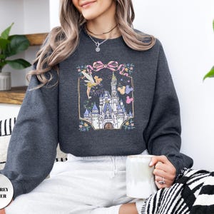 May include: Dark grey heather sweatshirt featuring a colorful graphic of a fairytale castle, Tinkerbell, and Mickey Mouse balloons. The design is framed by a pink bow and floral accents. The sweatshirt has a crew neck and long sleeves.
