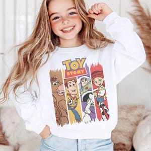 K&ouml;nnte beinhalten: Ein wei&szlig;es Langarm-Sweatshirt mit einem farbenfrohen Grafikdruck des Toy Story-Logos &uuml;ber vertikalen Feldern, die die Figuren Bullseye, Woody, Buzz Lightyear und Jessie zeigen. Das Sweatshirt hat einen Rundhalsausschnitt und gerippte B&uuml;ndchen.