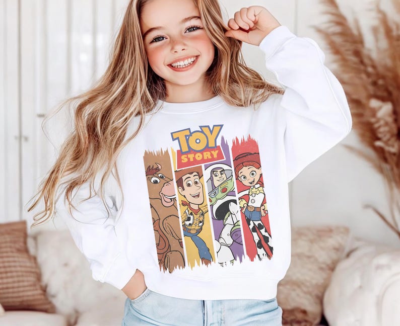 K&ouml;nnte beinhalten: Ein wei&szlig;es Langarm-Sweatshirt mit einem farbenfrohen Grafikdruck des Toy Story-Logos &uuml;ber vertikalen Feldern, die die Figuren Bullseye, Woody, Buzz Lightyear und Jessie zeigen. Das Sweatshirt hat einen Rundhalsausschnitt und gerippte B&uuml;ndchen.