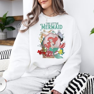 May include: A white long-sleeve sweatshirt featuring a colorful graphic print of The Little Mermaid characters including Ariel, Flounder, King Triton, and Ursula. The design includes underwater elements like a large seashell, coral, and bubbles.