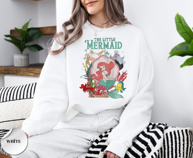 May include: A white long-sleeve sweatshirt featuring a colorful graphic print of The Little Mermaid characters including Ariel, Flounder, King Triton, and Ursula. The design includes underwater elements like a large seashell, coral, and bubbles.