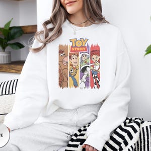 K&ouml;nnte beinhalten: Ein wei&szlig;es Langarm-Sweatshirt mit einem Grafikdruck des Toy Story-Logos &uuml;ber vier vertikalen Feldern, die die Figuren Bullseye, Woody, Buzz Lightyear und Jessie in ihren charakteristischen Farben und Outfits auf der Vorderseite zeigen.