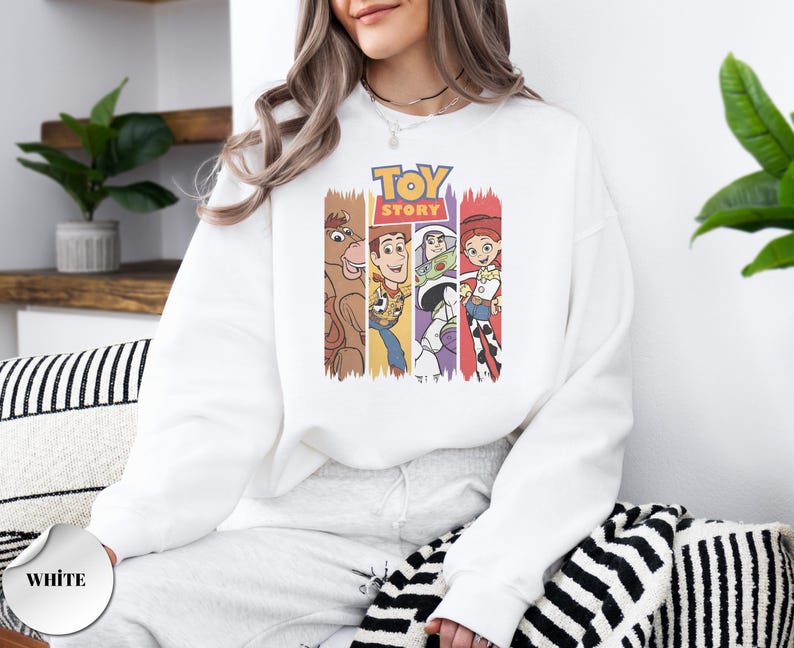K&ouml;nnte beinhalten: Ein wei&szlig;es Langarm-Sweatshirt mit einem Grafikdruck des Toy Story-Logos &uuml;ber vier vertikalen Feldern, die die Figuren Bullseye, Woody, Buzz Lightyear und Jessie in ihren charakteristischen Farben und Outfits auf der Vorderseite zeigen.