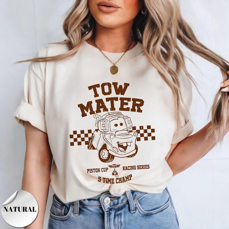 May include: A cream-colored short-sleeve t-shirt featuring a brown graphic of the character Tow Mater. The design includes a checkered flag pattern, the text TOW MATER, PISTON CUP RACING SERIES, and S TIME CHAMP, with a small trophy icon.