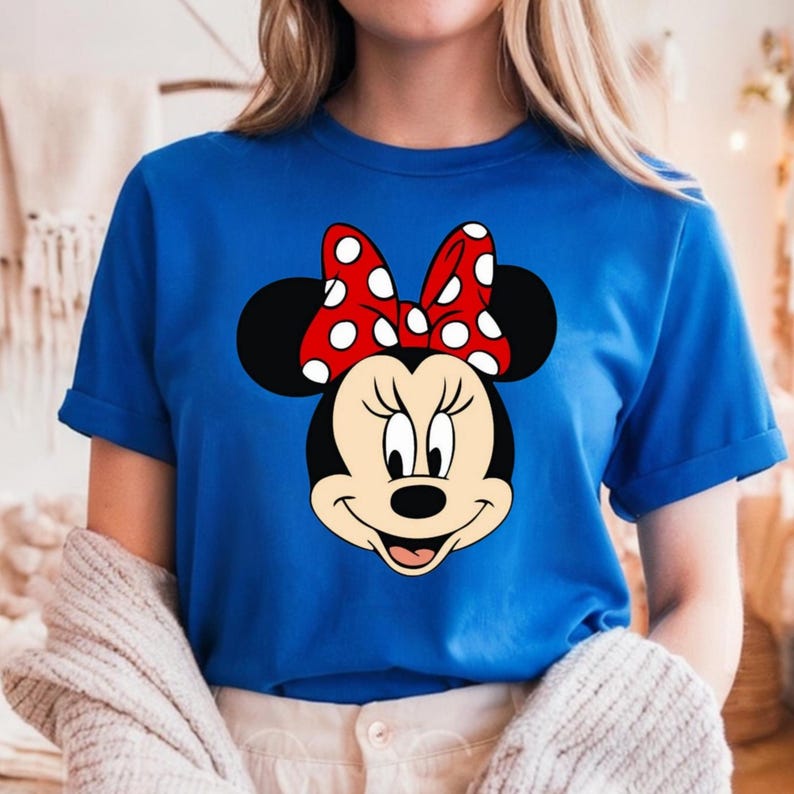 Puede incluir: Una camiseta azul real con un gr&aacute;fico de Minnie Mouse. Minnie Mouse se representa con orejas negras, una cara sonriente y un lazo rojo con lunares blancos. La camiseta tiene mangas cortas.