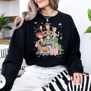 May include: A black crewneck sweatshirt featuring a colorful graphic print of various animated characters including a cowboy, space ranger, cowgirl, dinosaur, pig, potato figure, and others, with handwritten names like Forky, Jessie, and Hamm.