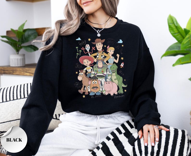 May include: A black crewneck sweatshirt featuring a colorful graphic print of various animated characters including a cowboy, space ranger, cowgirl, dinosaur, pig, potato figure, and others, with handwritten names like Forky, Jessie, and Hamm.