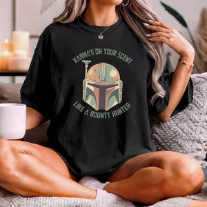 May include: A black short-sleeved t-shirt featuring a graphic of a Mandalorian-style helmet in muted green, orange, and brown tones. The text "KARMA'S ON YOUR SCENT LIKE A BOUNTY HUNTER" is printed in a circular arc around the helmet design.