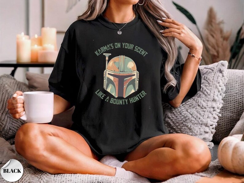 May include: A black short-sleeved t-shirt featuring a graphic of a Mandalorian-style helmet in muted green, orange, and brown tones. The text "KARMA'S ON YOUR SCENT LIKE A BOUNTY HUNTER" is printed in a circular arc around the helmet design.