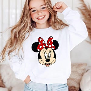 Puede incluir: Sudadera blanca con un dise&ntilde;o de Minnie Mouse. El gr&aacute;fico muestra la cara de Minnie con un lazo rojo con lunares blancos. La sudadera es de manga larga y tiene cuello redondo.
