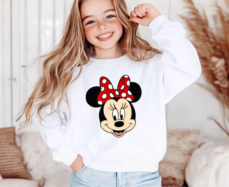 Puede incluir: Sudadera blanca con un dise&ntilde;o de Minnie Mouse. El gr&aacute;fico muestra la cara de Minnie con un lazo rojo con lunares blancos. La sudadera es de manga larga y tiene cuello redondo.