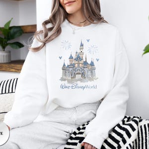 May include: A white crewneck sweatshirt featuring a graphic print of a castle with blue roofs and spires. Above the castle are blue fireworks and small Mickey Mouse head icons. The text Walt Disney World is printed in blue below the castle.