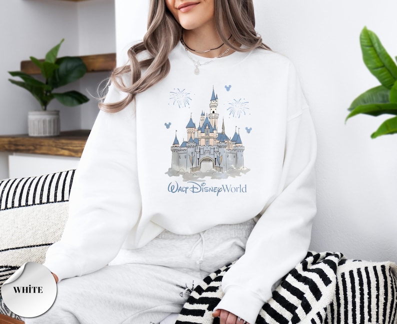 May include: A white crewneck sweatshirt featuring a graphic print of a castle with blue roofs and spires. Above the castle are blue fireworks and small Mickey Mouse head icons. The text Walt Disney World is printed in blue below the castle.