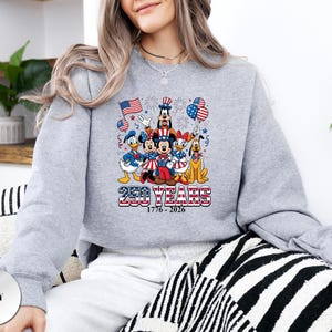 May include: A grey crewneck sweatshirt featuring a graphic of Disney characters including Mickey Mouse, Minnie Mouse, Donald Duck, Daisy Duck, Goofy, and Pluto. The design includes American flags, balloons, and the text 250 YEARS 1776 - 2026.