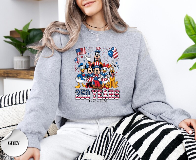 May include: A grey crewneck sweatshirt featuring a graphic of Disney characters including Mickey Mouse, Minnie Mouse, Donald Duck, Daisy Duck, Goofy, and Pluto. The design includes American flags, balloons, and the text 250 YEARS 1776 - 2026.