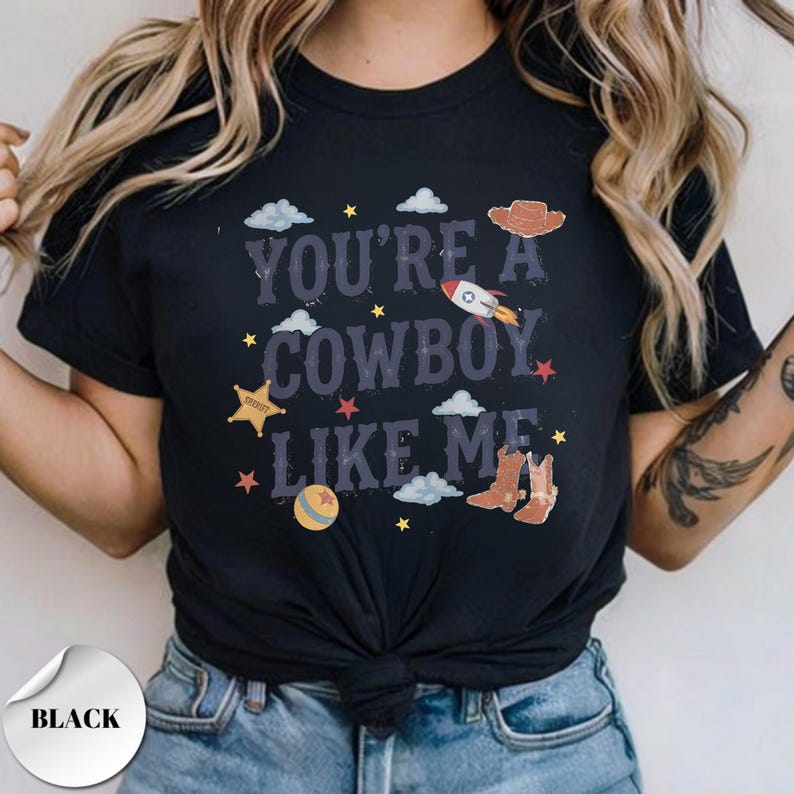May include: A black short-sleeved t-shirt featuring the text "YOU'RE A COWBOY LIKE ME" in a distressed font. The graphic includes illustrations of a brown cowboy hat, a rocket, a sheriff star, a ball, and cowboy boots, surrounded by small stars and clouds.
