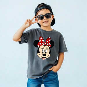 Op de afbeelding: Een grijs t-shirt met een Minnie Mouse-afbeelding, een rode strik met witte stippen. Het kind draagt een blauwe spijkerbroek, een zwarte pet en een zonnebril. De afbeelding toont een kind dat lacht en het vredesteken maakt.