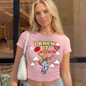 May include: A light pink short-sleeve t-shirt featuring a graphic print of a person playing a guitar, a red hat, white clouds, musical notes, and the text I KNEW IT I KNEW YOU TS inside a red heart. The shirt has a crew neckline and a fitted cut.