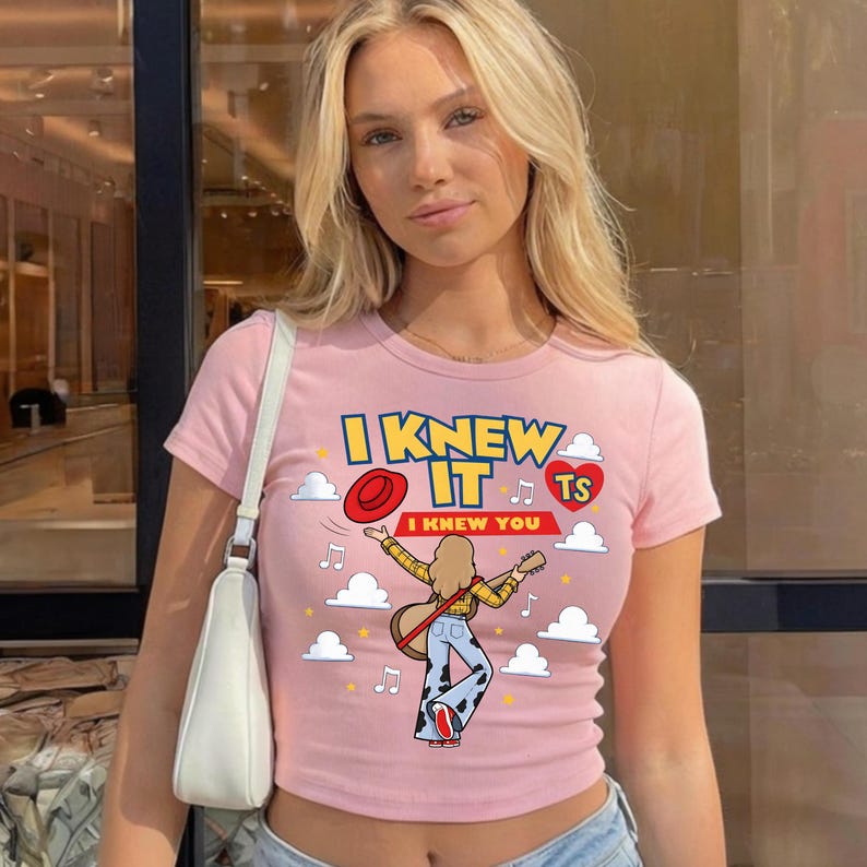 May include: A light pink short-sleeve t-shirt featuring a graphic print of a person playing a guitar, a red hat, white clouds, musical notes, and the text I KNEW IT I KNEW YOU TS inside a red heart. The shirt has a crew neckline and a fitted cut.
