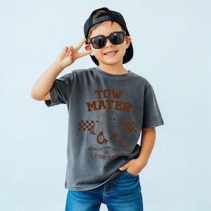May include: A charcoal gray short-sleeve t-shirt featuring a brown graphic of the character Tow Mater from the Piston Cup Racing Series. The design includes a checkered pattern, the text TOW MATER, and the words 5 TIME CHAMP.