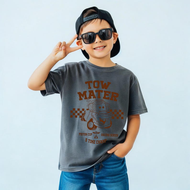 May include: A charcoal gray short-sleeve t-shirt featuring a brown graphic of the character Tow Mater from the Piston Cup Racing Series. The design includes a checkered pattern, the text TOW MATER, and the words 5 TIME CHAMP.