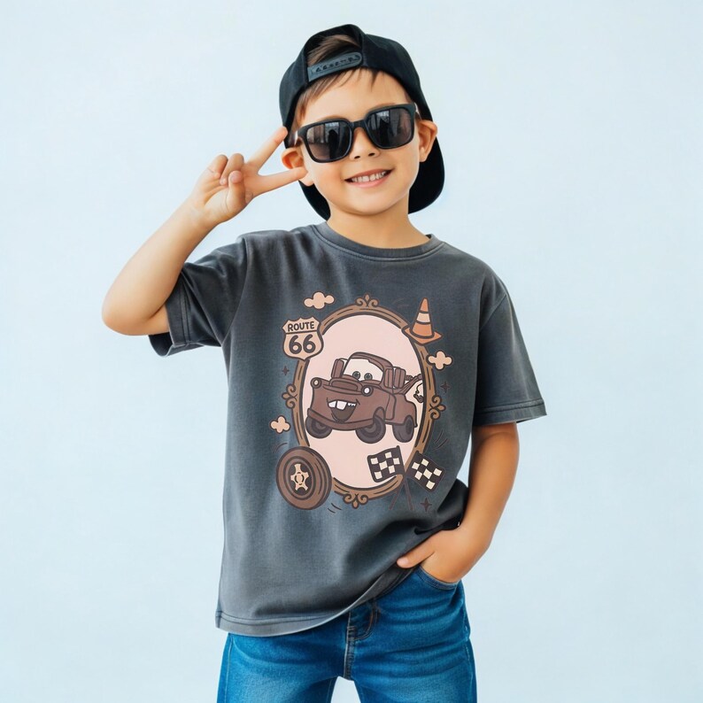 May include: A charcoal gray short-sleeve t-shirt featuring a graphic print of a brown tow truck character inside an oval frame. The design includes a Route 66 sign, a traffic cone, a tire, and checkered flags in shades of brown and cream.