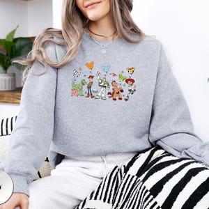 Op de afbeelding: Een grijs gem&ecirc;leerd sweatshirt met lange mouwen en een centrale grafische print van diverse geanimeerde speelgoedfiguren, waaronder een cowboy, een space ranger, een cowgirl, een dinosaurus en een varken, die ballonnen in muisorenvorm vasthouden.