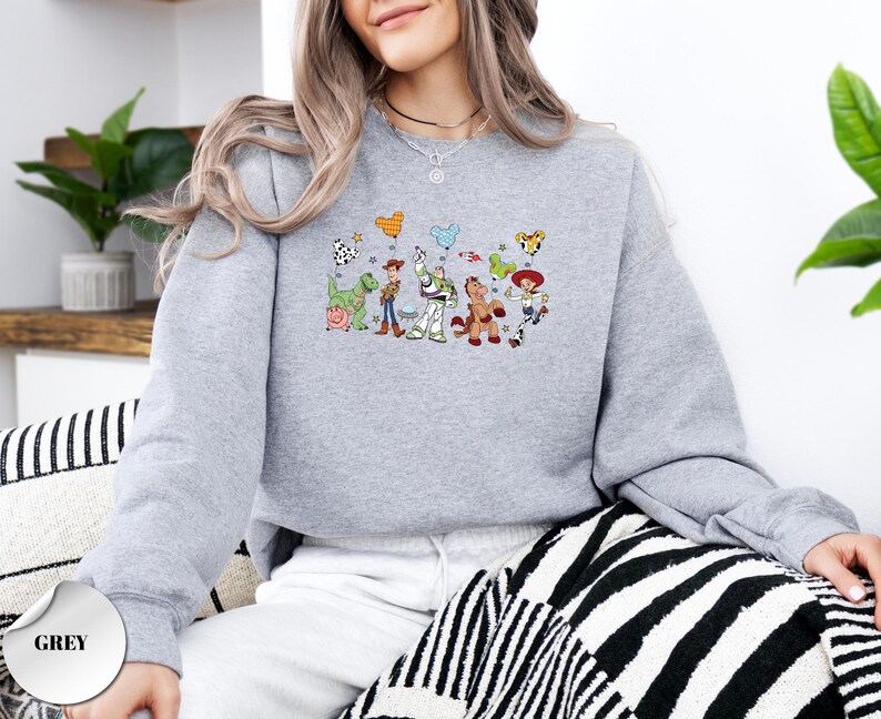 Op de afbeelding: Een grijs gem&ecirc;leerd sweatshirt met lange mouwen en een centrale grafische print van diverse geanimeerde speelgoedfiguren, waaronder een cowboy, een space ranger, een cowgirl, een dinosaurus en een varken, die ballonnen in muisorenvorm vasthouden.
