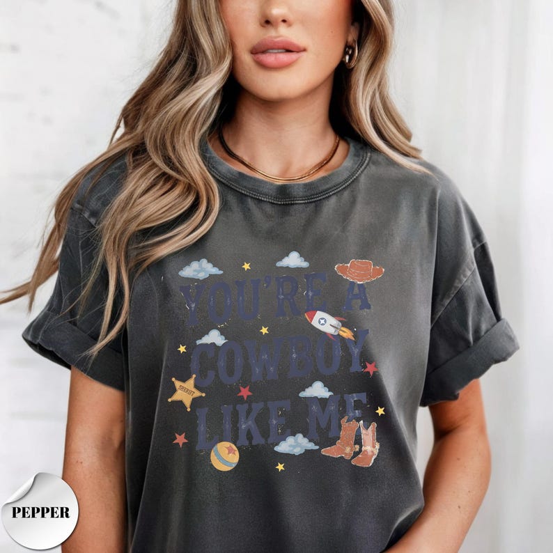 May include: A charcoal gray short-sleeve t-shirt featuring the text "YOU'RE A COWBOY LIKE ME" in dark blue letters. The graphic includes a brown cowboy hat, a rocket ship, a yellow sheriff star, a colorful ball, and brown cowboy boots.