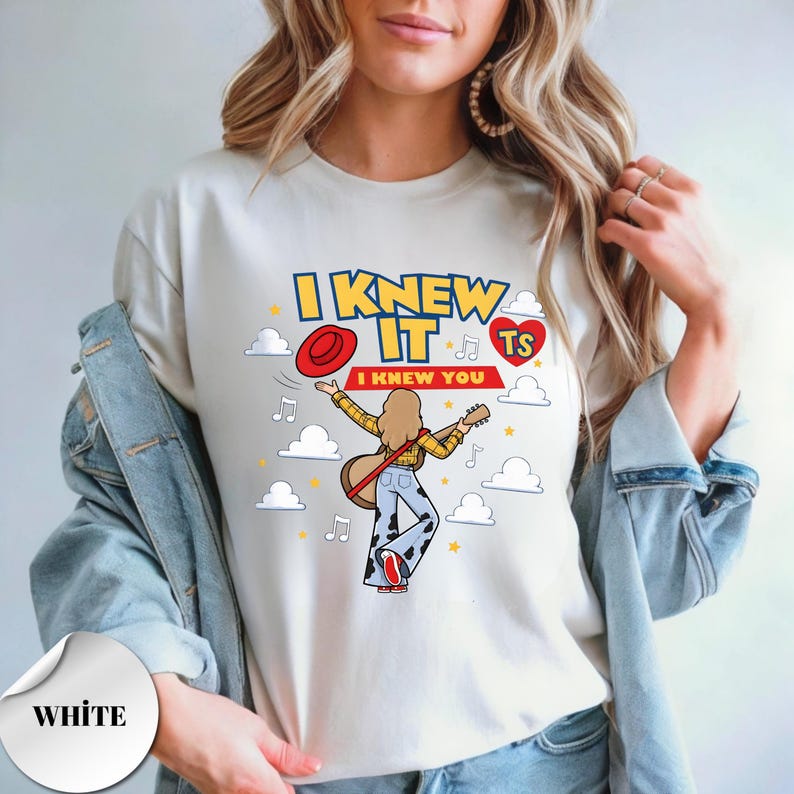 May include: A white t-shirt featuring a graphic of a person with long hair playing a guitar, wearing a yellow plaid shirt and cow-print pants. The design includes the text I KNEW IT and I KNEW YOU, a red hat, a red heart with TS, and white clouds.