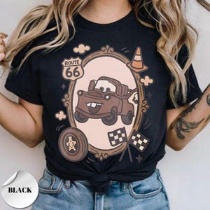 May include: A black t-shirt featuring a graphic of a brown tow truck inside an oval frame. The design includes a Route 66 sign, a traffic cone, a tire, and two checkered flags. The truck has large eyes on the windshield and a wide, toothy grin.
