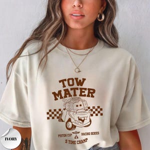 May include: An ivory short-sleeve t-shirt featuring a brown graphic print of the character Tow Mater. The design includes a checkered flag pattern, the text "TOW MATER", "PISTON CUP RACING SERIES", and "S TIME CHAMP" with a small trophy icon.