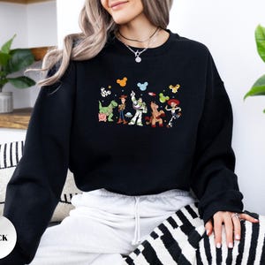 Op de afbeelding: Een zwart sweatshirt met lange mouwen en een kleurrijke grafische print van Toy Story-personages zoals Woody, Buzz Lightyear, Rex, Bullseye en Jessie, geaccentueerd met kleine Mickey Mouse-oorpictogrammen boven de figuren.