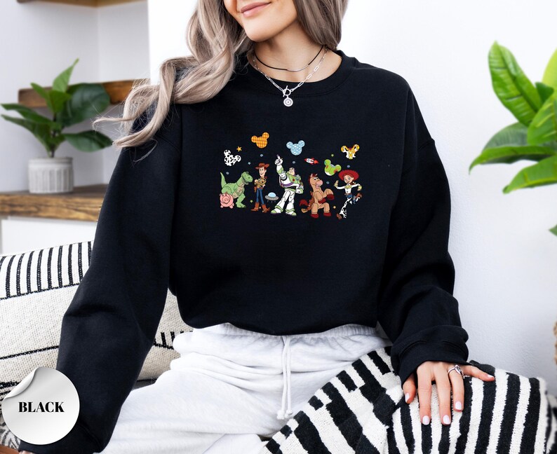 Op de afbeelding: Een zwart sweatshirt met lange mouwen en een kleurrijke grafische print van Toy Story-personages zoals Woody, Buzz Lightyear, Rex, Bullseye en Jessie, geaccentueerd met kleine Mickey Mouse-oorpictogrammen boven de figuren.