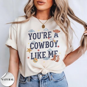 May include: A cream-colored short-sleeve t-shirt featuring the dark blue text "YOU'RE A COWBOY LIKE ME" surrounded by graphic illustrations of a rocket, a sheriff badge, cowboy boots, a hat, clouds, stars, and a ball in various colors.