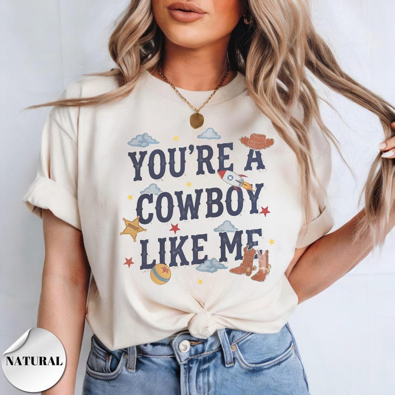 May include: A cream-colored short-sleeve t-shirt featuring the dark blue text "YOU'RE A COWBOY LIKE ME" surrounded by graphic illustrations of a rocket, a sheriff badge, cowboy boots, a hat, clouds, stars, and a ball in various colors.